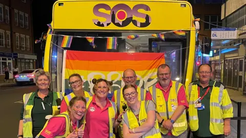 SOS BUS Volunteers and paid staff stood outside the SOS Bus. They are wearing polo shirts with yellow  hi-visibility vests and are smiling