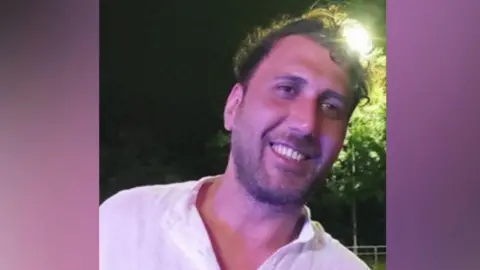 Mustafa Qays Obadey smiles while wearing a white shirt. He is posing at night with a street light seen shining behind his head.