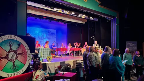 KIRSTY LAWRENCE People sat on a stage with microphones and a guitar with blue lighting behind them.