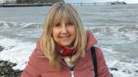 Supplied Ellen Cook wearing a pink coat and stood in front of a rocky coast. She has long light hair.