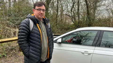Jamie Vaughan is wearing a blue and yellow puffer jacket, glasses and a grey polo shirt. He is stood next to his car in the Browne's Folly car park. He is looking at the camera.