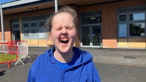 Photograph of Laneyle who is a student at Royal College Manchester. She is pictured in front of the Ged Mason building. She has swept-back light brown hair and is wearing a blue hoodie on top of a blue t-shirt.