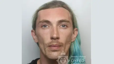 Police mugshot. The man has long blue and green hair.