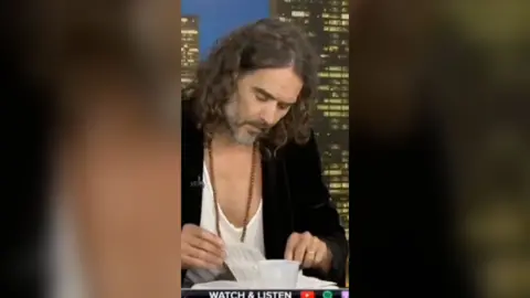 Russell Brand looks down at a Bible, flicking through the pages. He's wearing a black blazer and low-cut white t-shirt.