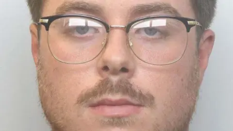 A man with short brown hair, a moustache and some facial hair and glasses is looking at the camera in a police mugshot.