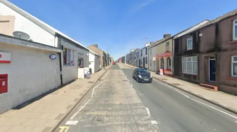 Google The A5086 is a single carriageway bordered by two-storey terraced houses. A post office and corner shop sign is on the left, there is a store front called: "Pasta Delight" on the left.