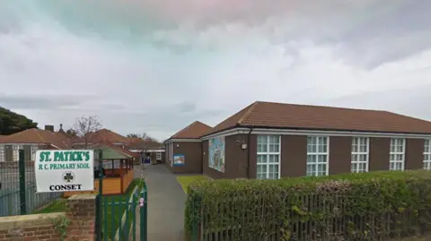 Brick school building behind gate and sign which reads in green: St Patrick's R C Primary School.