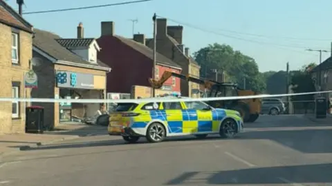 John Jest A yellow JCB is seen in the background of a police cordon. A police car is in the foreground immediately behind some police tape. In the background damage to the co-op can also be seen with a hole in the front of the store where the windows and cash machine previously were.