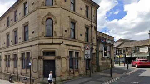 The exterior of a building on a street corner. It has three storeys and oblong windows. Some traffic lights and a pedestrian crossing stand next to it.