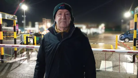 Michael looking at the camera. He is standing outside wearing a black coat and a beanie hat with unite embroidered on it.