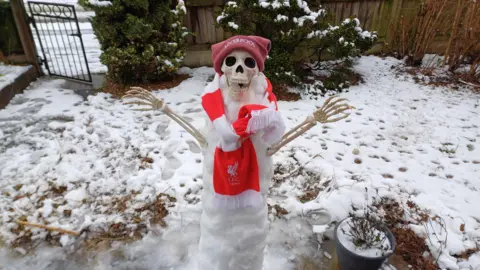 TheDevlins A snowman with a skeleton mask and arms wearing a Liverpool FC scarf and hat in a snow-covered garden.  