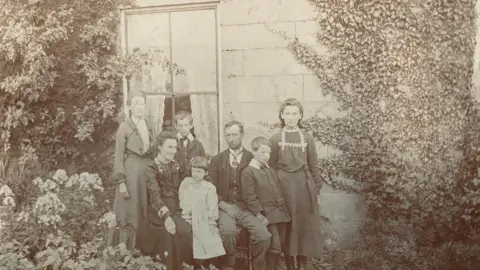 Martin Beal A black and white picture of a family group sitting and standing in front of a window. It looks to date from the very early twentieth century.
