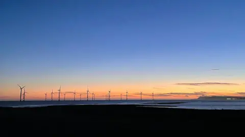 BBC Weather Watchers/Superted A sunrise or sunset over the coastline. Wind turbines sit out to sea with the sky orange around them with wispy clouds. The sky further up is blue. The ground in front of the sea is completely black and the sea looks dark.