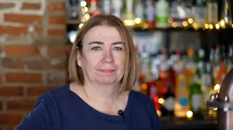Jamie Niblock/BBC Tina Leamon with shoulder length hair and a blue top standing behind a bar. Bottles can be seen in the background.