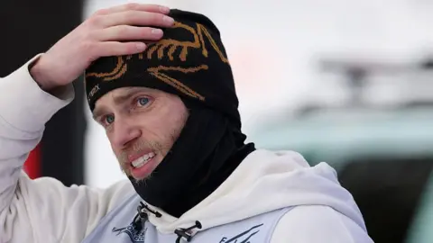 Gus Kenworthy is wearing a black beanie and snood and has his hand on his head
