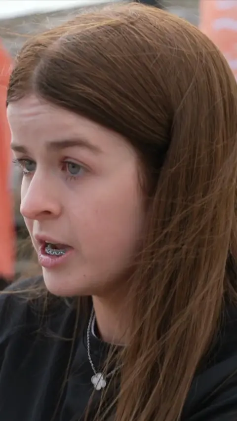 A woman with long brown hair wearing a black t-shirt and silver necklace. 