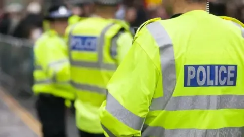 The back of a police officer who is wearing a yellow hi-vis jacket with silver reflective stripes. A logo that says "police" is on a blue background. In the background of the picture, two other officers are wearing the same jackets, as well as black police hats.