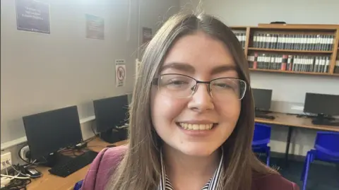 Aleigha, a teenage girl, with glasses, a claret blazer and striped blue and white blouse. There are computers on a table in the background.