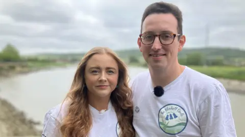 Alyce and Rob Le Brunn-Healey in matching white t-shirts, smiling for the camera. A river is visible behind them.