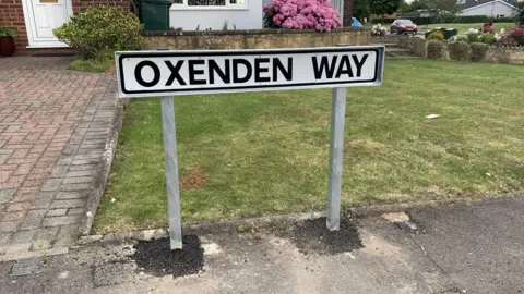 The 'Oxenden Way' sign standing on a pavement next to grass by a house