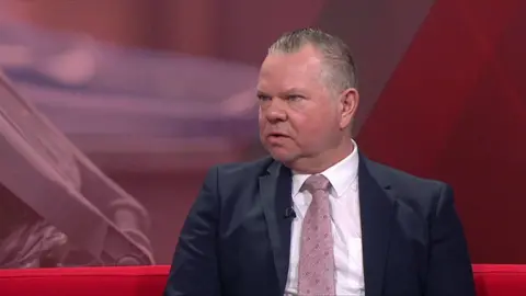A man wearing a suit and pink tie sits on red sofa looking at the camera. A screen can be seen behind him.