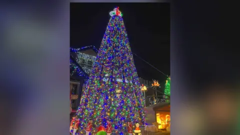 Paul Molyneux A towering Christmas tree wrapped in thousands of multicolored lights stands in front of the house. The tree is topped with a glowing ornament and surrounded by smaller decorations. The vibrant colors create a dazzling centerpiece.