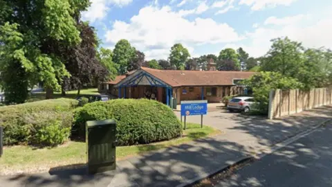 A single storey building with a car park, bordered by small hedges. A sign saying 'Mill Lodge' is at the entrance of the car park.
