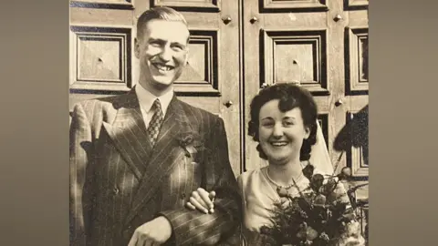 Contributed A black and white image of a young man and woman on their wedding day. They stand in front of a large wooden door and smile at the camera. The man has short hair and wears a suit, tie and shirt. The woman wears a white wedding dress and holds a bouquet of flowers in one hand while her other hand rests on the man's forearm. 