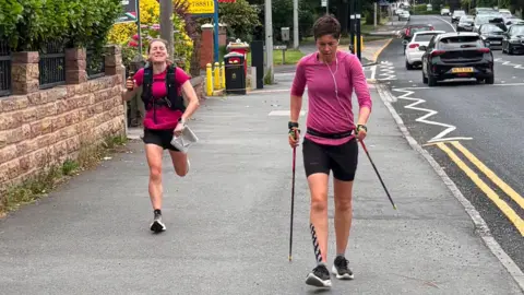 Zoe Murphy Trish Patterson, wearing a pink top and black shorts and shoes, on the right with walking sticks and walking along a road with cars travelling along. She has headphone in and is focused on the floor. She has tap on the front of her leg.