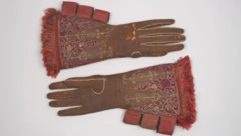 A pair of large brown leather gloves with red silk embroidery and gold embellishment, belonging to King James I, circa 1610s. They are on a grey background.