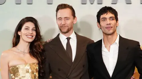 The trio stand with their hands around each other against a white background. Morrone wears a gold strapless top, her dark hair loose over one shoulder. Hiddleston wears a brown suit jacket and tie, with white shirt, he has a short goatee beard and short dark hair. Calva wears a black suit which is closed with one button, and a white shirt which is open at the collar. He is wearing small hoop earrings and has short dark hair. 