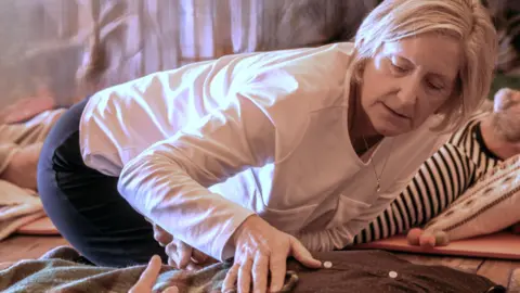 A close-up of Lyndsey, who is in a room with a couple of people lying on the floor on yoga mats. She is leaning over next to one client, with her hand laid gently on their chest.