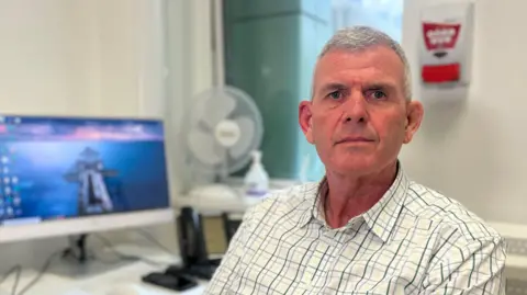 Simon sits in an office beside a desk with two computer monitors, a keyboard, and various desk items. A fan and other equipment are in the background, giving the space a clinical or administrative feel.