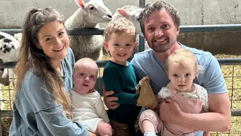 The Strange family. The parents are crouched down and are holding their younger children in their arms, while the older son stands between them. They are in a farm with sheep behind them