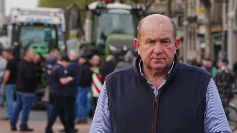 PA Media A man looks toward the camera. He is wearing a blue and white shirt, and a navy-blue gilet jacket. Behind him, out of focus, are two green tractors in an urban area. 