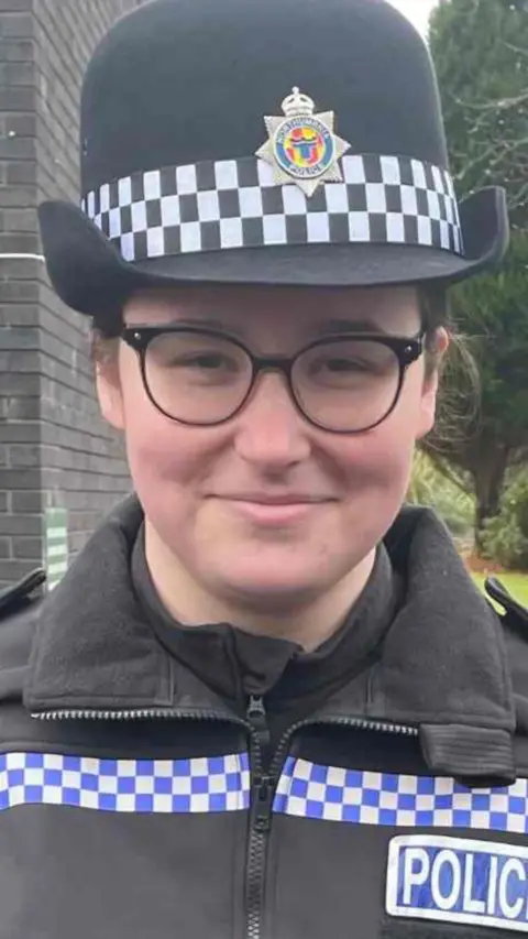 Jess Turnbull, wearing police uniform and smiling. She is standing in front of a dark grey brick building with a wooden entrance to one side. Behind her to the other side is a patch of grass, a tall, thin bush-like tree and some other trees, like you might find in the gardens of an office building. Jess is wearing a rounded police hat and black rimmed glasses and has her dark hair tied back.