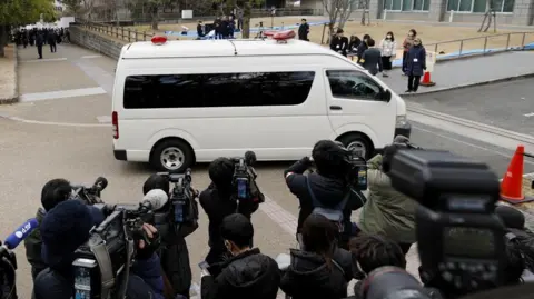 Reuters Een wit politiebusje met daarin Tetsuya Yamagami arriveert bij een districtsrechtbank in Nara, voorafgaand aan zijn hoorzitting over de veroordeling. Journalisten en videografen verdringen zich eromheen