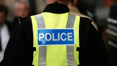 A generic shot of a police officer from behind. They are wearing a hi-viz vest with the word 'police' printed on the back. 