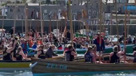 A larghe number of people in wooden rowing boats with oars held upwards with a harbour behind.