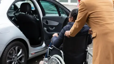 A wheelchair user with black hair and glasses - wearing a black puffer jacket and jeans - being assisted by a parent or carer wearing a camel-colored coat next to a silver car with one back door open. 