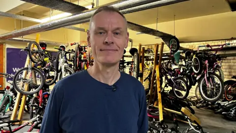 Alex Simons, wearing a blue jumper, stands in a workshop full of bicycles.