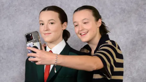 Madame Tussauds London Bella Ramsey wearing a black and cream hooped top stood behind a waxwork of them. They have a phone in their hand and are taking a selfie with the figure.