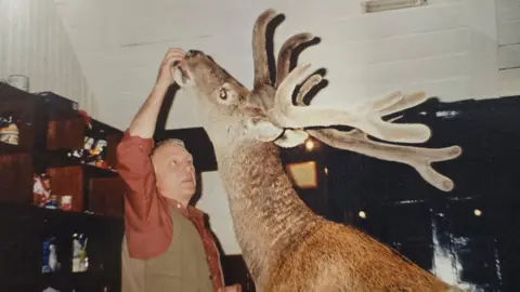 Hostelling Scotland A man feeds a stag inside the hostel. The stag, which has large antlers, is raising its head to reach the food.