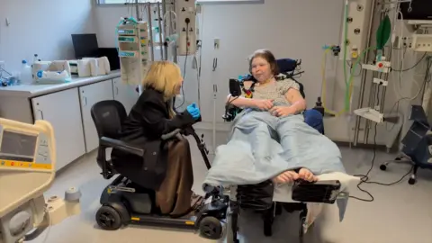 Dan Nelson / BBC Lucinda Ritchie in a hospital ward with BBC correspondent Nikki Fox. Lucinda is reclined on a chair with feet supported, while Nikki sits nearby in her powerchair, leaning forward. Surrounding equipment includes monitors, tubing, cabinets, and clinical supplies, indicating a healthcare environment.
