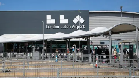 Getty Images An entrance to London Luton Airport. It is a grey building with London Luton Airport written in white on its wall. In front of it are barriers and a white covered walkway. People can be seen walking along