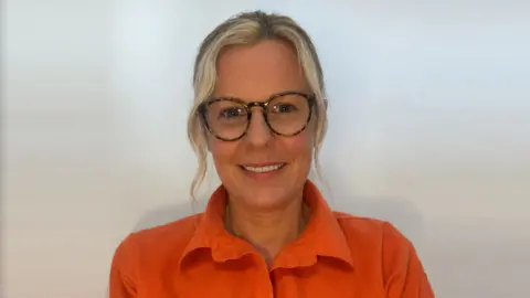 Glynis Sheppard Swimming teacher Glynis Sheppard is smiling towards the camera. She has blonde hair which is tied up with some strands coming down and is wearing glasses. She is wearing an orange blouse. 