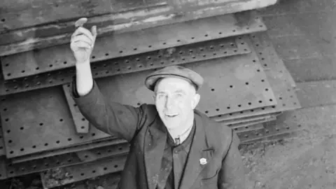 A black-and-white photograph of a worker in the Swan Hunter Shipyard on Tyneside on 8 January, 1940. An iron shipwright is shouting directions up to workers in the scaffolding. There are panels on the floor.