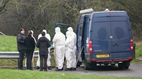 Ant Saddington/BBC A group of forensics officers dressed in all white with their faces covered standing in front of the side entrance to a navy blue transit van. Next to them are three other people dressed in all black.