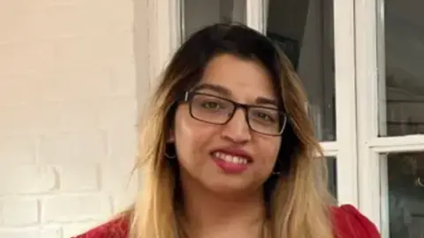 A close-up image of Lowestoft Mayor Nasima Begum. She is wearing glasses and a red t-shirt and is standing in front of a window. She is smiling into the camera.