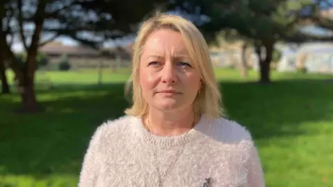 Caroline Court, wearing a white fluffy jumper with a broach and a necklace, looking straight at the camera. Her blonde hair falls to her shoulders. Behind her are trees and grass, which are out of focus. It is a bright, sunny day.